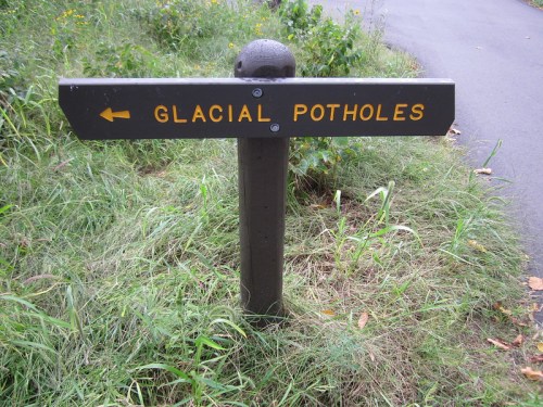 A wooden signpost with an arrow pointing left, labeled "GLACIAL POTHOLES," surrounded by grass and a pathway.
