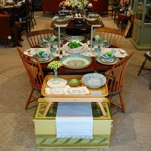 A beautifully arranged dining table with vintage dishes, greenery, and decorative items in a cozy shop setting.