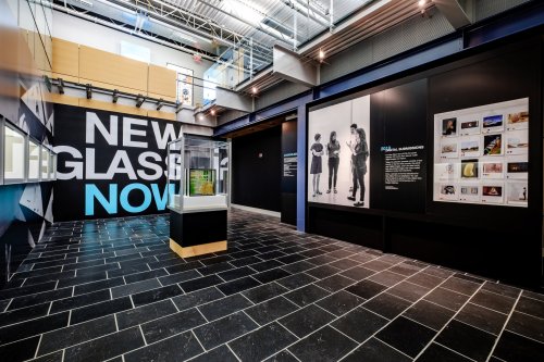 Interior view of an exhibition space featuring a display case and large wall text reading "New Glass Now."