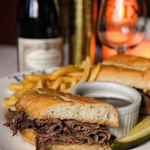A close-up of a roast beef sandwich with fries and dipping sauce, accompanied by a glass of wine in the background.