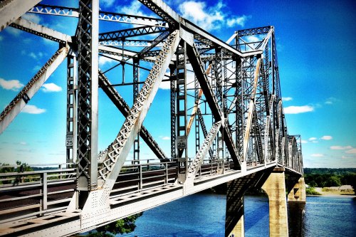 A metal truss bridge spans a river under a bright blue sky with scattered clouds.