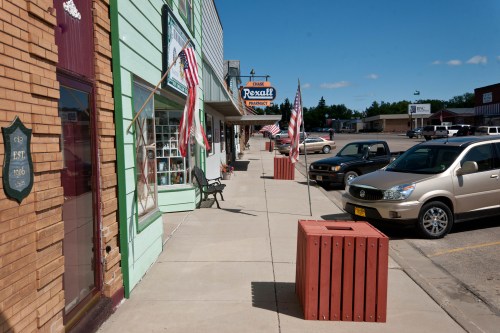 A small-town street with storefronts, American flags, and parked cars under a clear blue sky.
