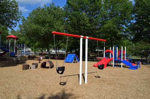 A colorful playground featuring swings, slides, climbing structures, and shaded trees on a sunny day.