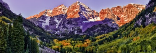 Majestic mountains with snow-capped peaks rise above a vibrant valley of autumn foliage and evergreen trees.