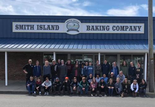 A large group of people poses in front of the Smith Island Baking Company building on a sunny day.