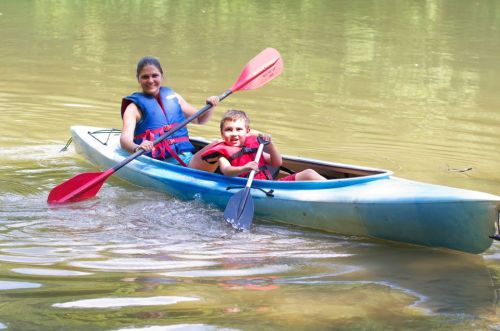 A woman and a boy paddle a kayak on calm water, both wearing life jackets and smiling.