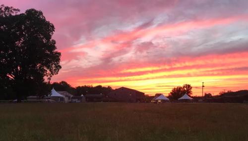 A vibrant sunset paints the sky in shades of pink and orange over a rural landscape with tents and trees.