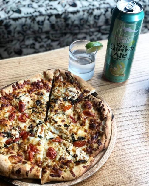 A pizza with toppings on a wooden plate, next to a can of Sea Quench Ale and a glass of water with a lime wedge.