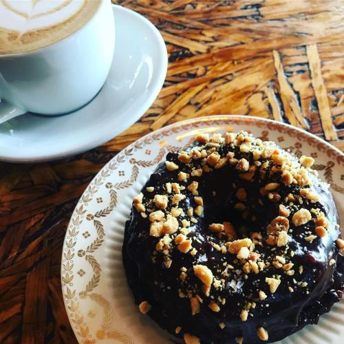 A chocolate donut topped with crushed nuts, next to a cup of latte on a wooden table.
