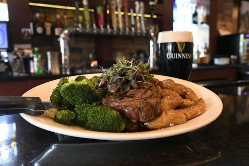 A plate of steak with broccoli and potatoes, alongside a pint of Guinness beer at a bar.