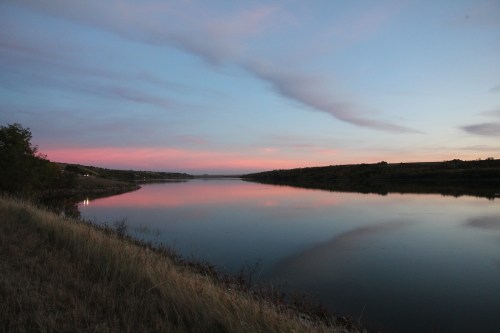 A serene river at dusk, reflecting soft pink and blue hues in the sky, surrounded by lush greenery.