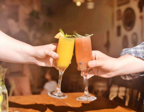 Two hands clink glasses of colorful cocktails, one yellow and one pink, in a warm, inviting setting.