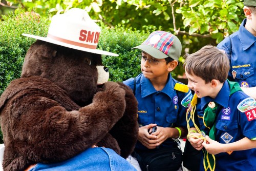 A bear mascot in a hat interacts with two young boys in scout uniforms, smiling and enjoying the moment together.