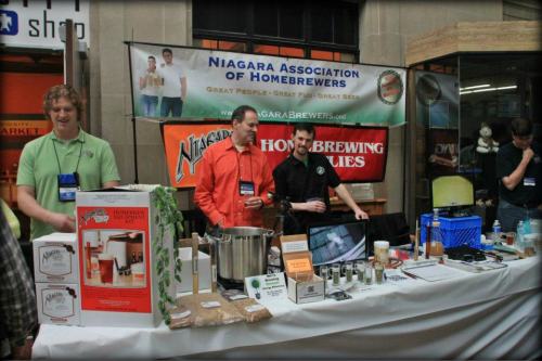 A booth at a homebrewing event with people showcasing brewing equipment and products, featuring a banner for the Niagara Association of Homebrewers.
