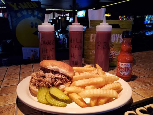 A plate with a pulled pork sandwich, crinkle fries, pickles, and three bottles of BBQ sauce on a table in a diner.