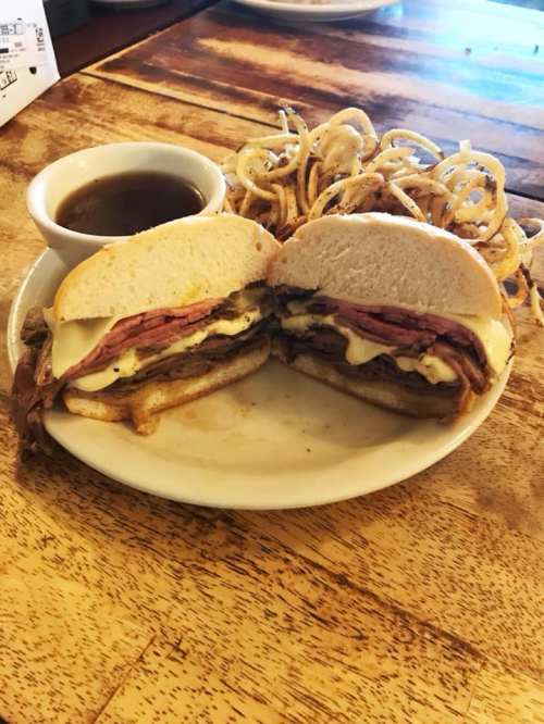 A sandwich filled with layers of meat and cheese, served with curly fries and a small cup of dipping sauce.
