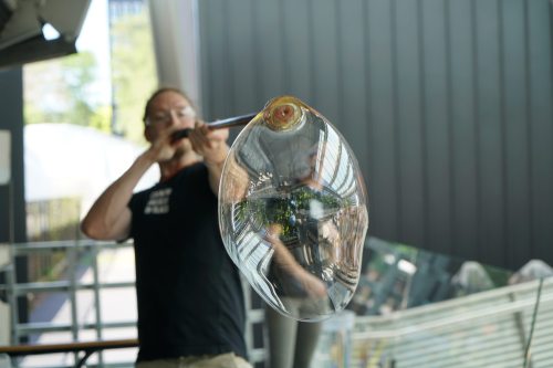 A glassblower shapes a large glass bubble while blowing into a pipe, with reflections visible in the glass.