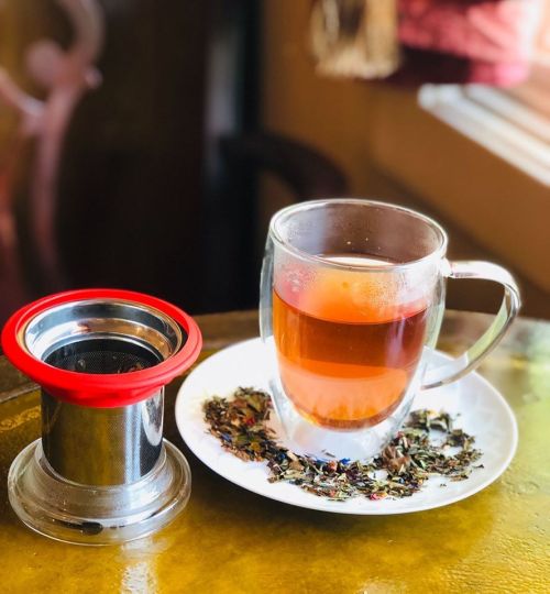 A glass cup of tea on a plate, with loose tea leaves scattered around and a tea infuser beside it.