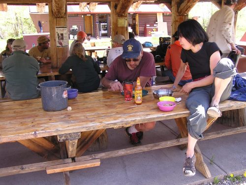 A group of people sitting at a wooden picnic table, with two individuals engaged in conversation and food preparation.