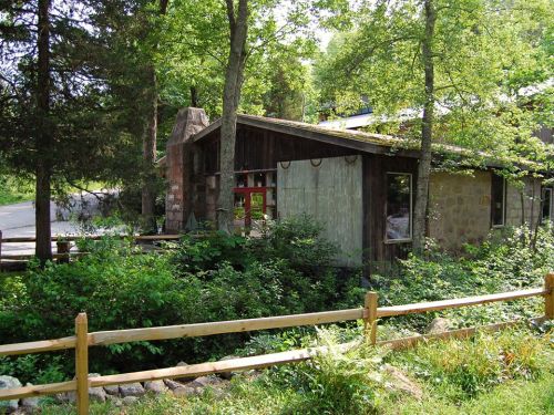 A rustic stone house surrounded by trees and greenery, with a wooden fence in the foreground.