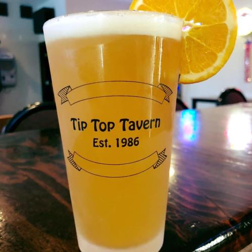 A pint glass of beer with an orange slice, featuring the logo of Tip Top Tavern, established 1986.