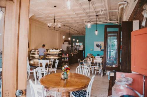 Cozy café interior with wooden tables, white chairs, and a display case, featuring a blue accent wall and floral decor.