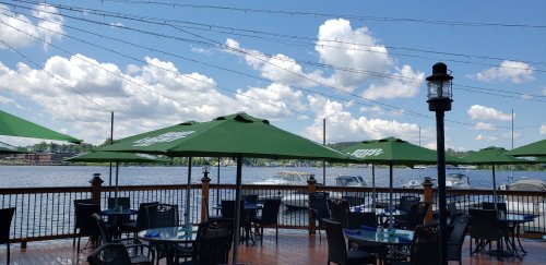 Outdoor dining area by a lake, featuring green umbrellas, tables, and boats in the water under a blue sky with clouds.
