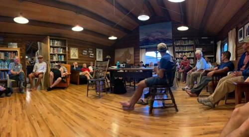 A group of people seated in a cozy, wooden room filled with books, engaged in conversation or discussion.