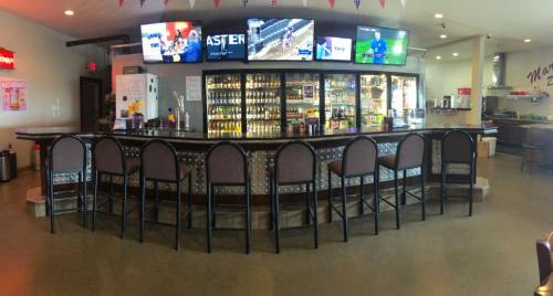A bar with high stools, multiple TVs displaying sports, and a well-stocked drink fridge in a casual setting.