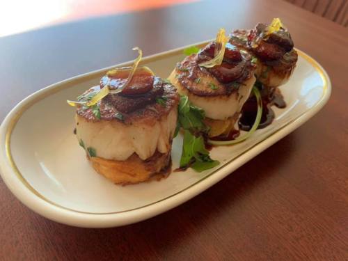Three beautifully plated scallops topped with herbs and garnishes, served on a white dish with a drizzle of sauce.