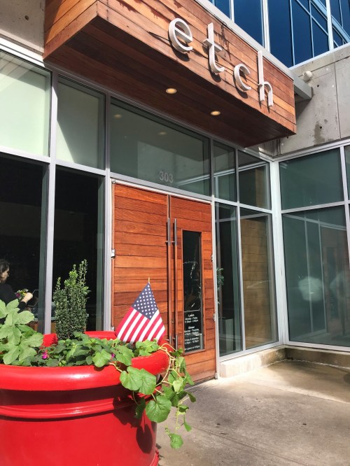 Exterior of a restaurant named "etch," featuring a wooden sign and a red planter with a small American flag.