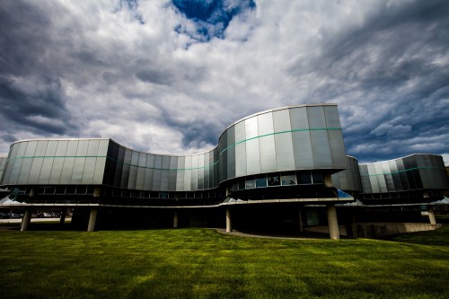 Modern building with a curved design, large glass windows, and a grassy foreground under a cloudy sky.