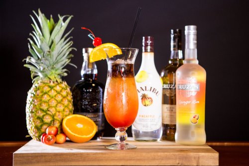 A colorful cocktail garnished with fruit, surrounded by bottles of rum and a fresh pineapple on a wooden surface.