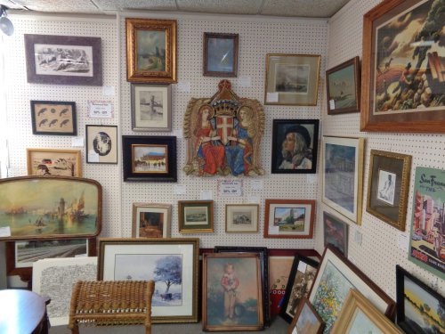A wall display of various framed artworks and photographs in an antique shop, featuring diverse styles and subjects.