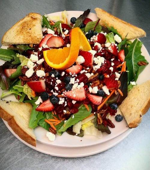 A colorful salad with mixed greens, strawberries, blueberries, orange slices, pecans, feta cheese, and toasted bread.