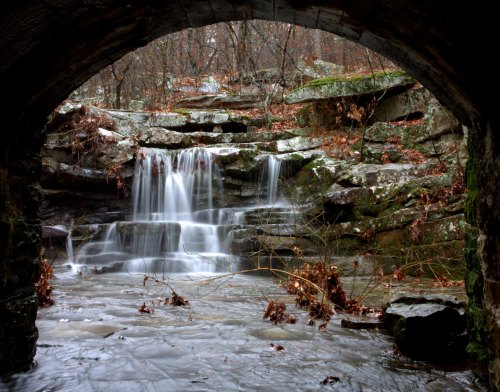 A serene waterfall cascades over rocks, framed by an archway, surrounded by autumn leaves and a tranquil stream.