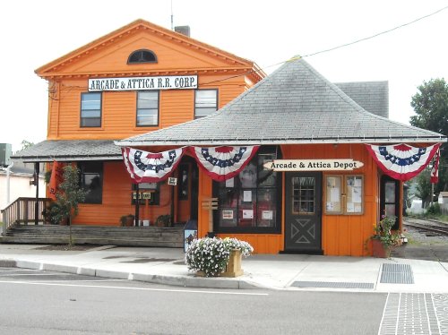 Historic orange train depot with decorative bunting, featuring a sign for Arcade & Attica R.R. Corp. and a flower planter.