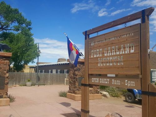 Sign for Fort Garland Museum with Colorado flag, surrounded by a rustic landscape under a blue sky. Open daily 9am-5pm.