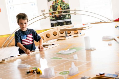 A young boy in a flag shirt plays with a model city layout, moving toy vehicles on a large map.