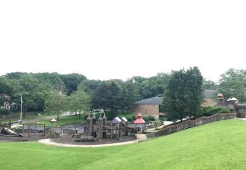 A playground with wooden structures and colorful roofs, surrounded by green grass and trees, under a cloudy sky.