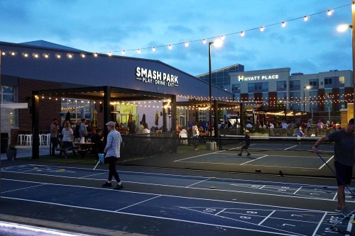 Outdoor entertainment venue with string lights, people socializing, and a nearby hotel in the background.
