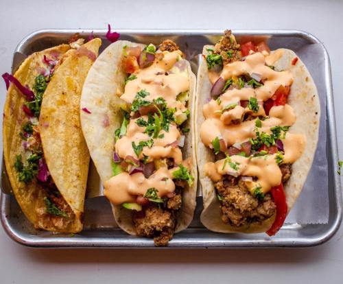 Three delicious tacos on a metal tray, filled with fried chicken, fresh vegetables, and drizzled with a creamy sauce.
