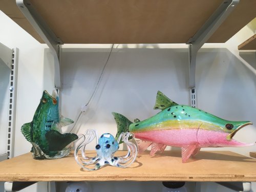 Three colorful glass sculptures: a green fish, a pink fish, and a blue octopus, displayed on a wooden shelf.