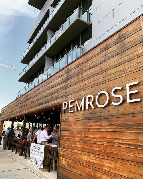 Exterior of a modern building with a wooden facade, featuring the name "PEMROSE" and a bustling outdoor dining area.