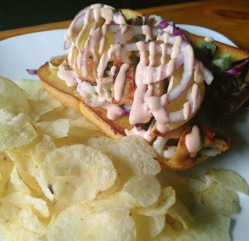 A sandwich topped with sliced pickles and sauce, served with a side of potato chips on a white plate.