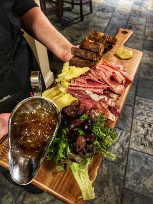 A wooden platter with assorted meats, cheeses, olives, greens, and a small bowl of jelly, served by a person.