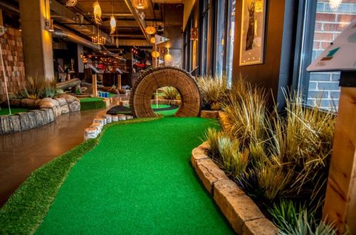 A vibrant indoor mini-golf course featuring a green carpet, decorative rocks, and a wooden tunnel.