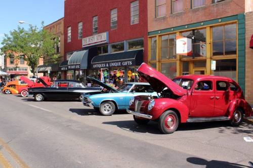 A row of classic cars parked on a street, with hoods open, in front of a colorful antique shop.