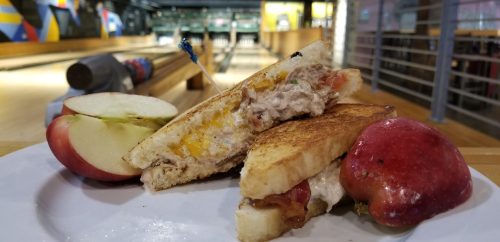 A plate with a grilled sandwich and apple slices, set against a bowling alley backdrop.