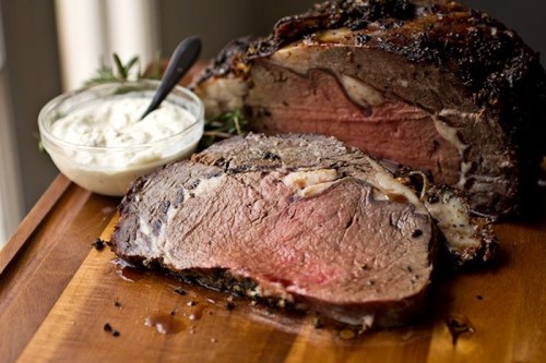 A succulent roast beef on a wooden cutting board, with a bowl of creamy sauce and herbs beside it.